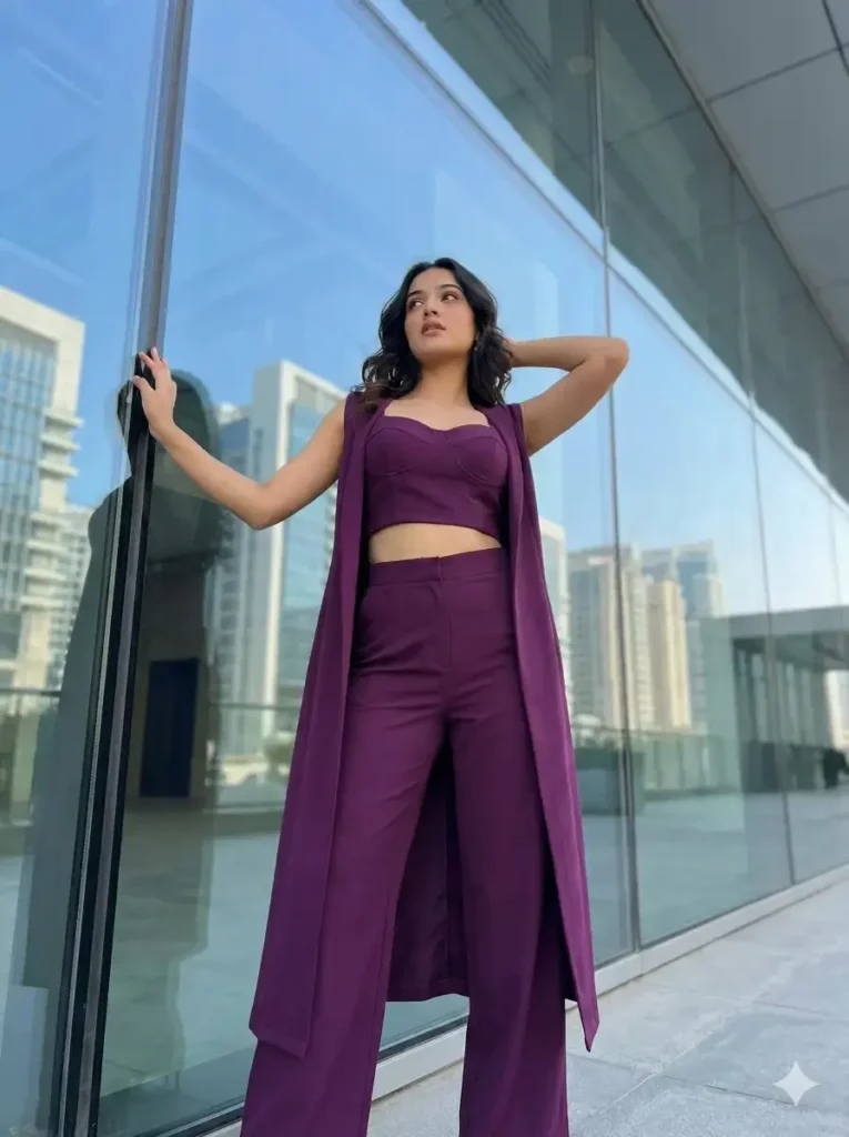 11 Gemini Prompts for Stunning Women Photos Create a contemporary fashion portrait of a young woman standing confidently against a reflective glass wall in a modern architectural setting. She wears a rich, deep-purple coordinated outfit consisting of a structured crop top, high-waisted wide-leg pants, and a long, sleeveless open-front overlay that flows vertically along her silhouette. The fabric has a subtle texture and matte sheen, adding depth without distraction. Her pose is bold and expressive: one arm extended outward, lightly touching the glass, while the other is bent with her hand resting behind her head. This creates a strong, asymmetrical composition that emphasizes confidence and elegance. Her hair is styled in soft, loose waves cascading over one shoulder. Makeup is modern. She gazes slightly upward and away from the camera with a calm, self-assured expression. Use aspect ratio 3:4.